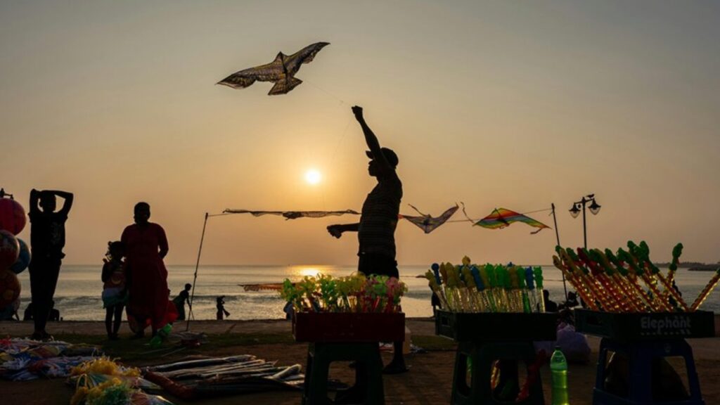A man silhouetted against a low sun flies a kite on a Sri Lankan seafront, beside stalls of brightly coloured toys and balloons, with the sea behind him
